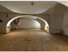 Gewölbter Kellerraum mit weiß gekalkten Backsteinwänden und zwei großen Rundbögen, teilweise mit freiliegendem Ziegelmauerwerk. Ziegelboden, einfache Deckenleuchte sowie sichtbare Leitungen und ein kleiner Wandschrank an der rechten Seite.