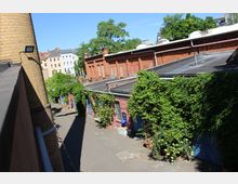 Innenhof eines ehemaligen Industrieareals mit niedrigen Backsteingebäuden und Flachdächern, teils mit Efeu und dichter Begrünung überwachsen. Links steht ein hoher runder Schornstein, im Hintergrund sind Wohnhäuser und große Bäume zu sehen.