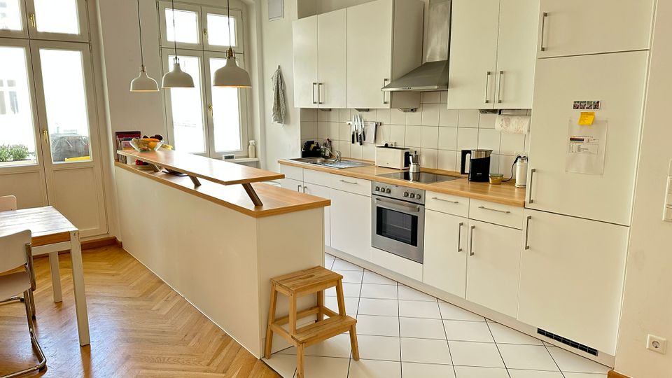 Helle, moderne Wohnküche mit weißen Schränken, Holzarbeitsplatten und einer langen Kücheninsel mit schmaler Ablage. Große Fenster und drei Hängelampen beleuchten den Raum; sichtbar sind Herd mit Edelstahl-Dunstabzug, Spüle sowie ein Mix aus Fliesenboden im Küchenbereich und Fischgrätparkett im Essbereich.