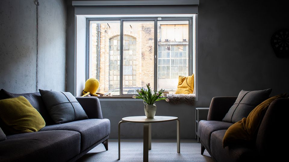 Modernes Wohnzimmer im Industriestil mit zwei grauen Sofas, gelben Kissen und einem runden Couchtisch mit Vase in der Mitte. Großes Fenster mit breiter Fensterbank und Kissen, Blick auf ein Backsteingebäude mit hohen Sprossenfenstern.