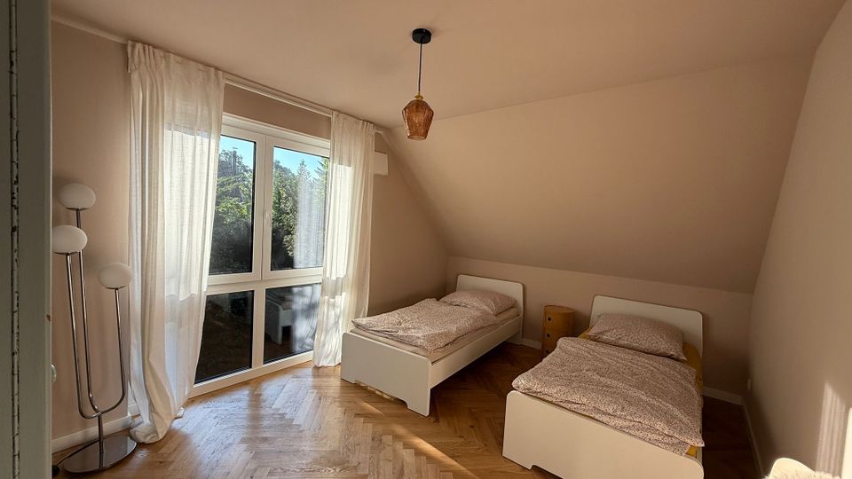 Schlafzimmer mit zwei einzelstehenden Betten und Holzfußboden in Fischgrätmuster. Ein großes Fenster mit lichtdurchlässigen Vorhängen bietet Blick auf eine grüne Außenumgebung, und eine Stehlampe steht in der Ecke.
