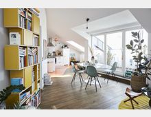 Helle Wohnung in einem Dachgeschoss, bestehend aus einem Wohn- und Essbereich mit skandinavischem Design. Zu sehen sind eine gelbe Bücherregalwand, ein Esstisch mit grünen Stühlen, große Fenster mit Zugang zur Terrasse und eine angrenzende Küche mit weißen Möbeln und offenen Dachfenstern.