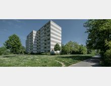 Wohnblock mit mehreren Etagen und abwechselnden horizontalen Streifen aus hellem und dunklem Beton, umgeben von einer Grünfläche mit Bäumen und einem Gehweg auf der rechten Seite, der in eine bewaldete Umgebung führt.