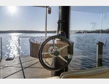 Steuerstand eines Bootes mit einem polierten Metall-Steuerrad und Holzkonsole, aufgenommen vor einer gläsernen Kabinenwand mit Blick auf einen ruhigen See unter klarem Himmel.