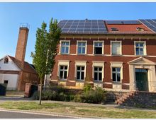 Backsteinhaus mit weißen Fensterrahmen und Sprossenfenstern, auf dem Dach sind Solarpaneele installiert. Links daneben steht ein kleineres Gebäude mit Ziegeldach und einem hohen Backsteinkamin, davor ein gepflasterter Weg und ein Baum.