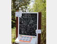 Tafel mit Kreidezeichnungen und Schriftzügen, darunter eine kleine Ablage mit Gästebuch und Schildern, die "Ahoi und herzlich willkommen" sowie "Gästebuch" anzeigen; die Szene befindet sich in einer grünen, bewaldeten Umgebung.