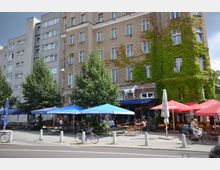 Mehrstöckiges Wohngebäude mit hellbrauner Fassade, teilweise mit grüner Kletterpflanze bedeckt. Im Erdgeschoss befinden sich Cafés oder Restaurants mit blau-roten Sonnenschirmen, Außenbestuhlung und Fahrradstellplätzen entlang der Straße.