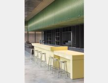 Moderne Bar mit gelber Tresenfront und silbernen Barhockern in einem Innenraum mit grauem Fußboden. Oberhalb der Bar befindet sich eine großflächige grüne Deckenverkleidung, dahinter dunkle Regale und eine Metallwand.