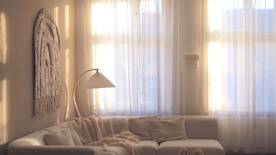 Sunlit living room with tall windows covered by sheer curtains, an L-shaped white sectional sofa, and a low round wooden coffee table on a fluffy rug. Neutral decor includes a wall hanging, an arched floor lamp, a sculptural ceiling light, and wooden flooring.