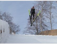 Ein Skispringer in schwarzer Ausrüstung und mit grünen Skiern fliegt durch die Luft über eine schneebedeckte Sprungschanze mit kahlen Bäumen im Hintergrund.