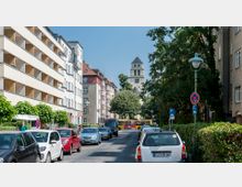 Straßenszene in einem Wohngebiet mit geparkten Autos auf beiden Seiten der Straße, mehrstöckigen Wohngebäuden links und rechts, sowie einer Kirche mit Turm und Uhr im Hintergrund. Im Vordergrund befinden sich Straßenlaternen und Verkehrszeichen, mit Bäumen, die den Gehweg flankieren.