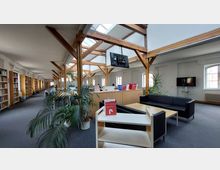 Offener Lesesaal einer Bibliothek mit Holzbalkenkonstruktion und Oberlichtern. Links stehen Bücherregale, in der Mitte befinden sich Sitzmöglichkeiten und Grünpflanzen, rechts eine kleine Sitzecke mit Sofas und einem Couchtisch.