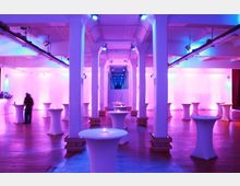 Großer Veranstaltungsraum mit hohen weißen Säulen, Holzfußboden und farbiger Beleuchtung in Pink und Blau. Im Raum stehen mehrere mit Stoff überzogene Stehtische, die teils mit Kerzen dekoriert sind.