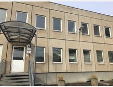 Zweistöckiges, schlichtes Büro- oder Verwaltungsgebäude mit beigefarbener Betonfassade und Reihen rechteckiger Fenster. Links ein Eingang mit Treppe und gebogenem Glasvordach; daneben ein Schild „K Haus 1“ und eine Außenleuchte an der Fassade.