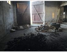 Alter, verlassen wirkender Innenraum mit offenen Holztoren, die nach außen führen. Auf dem Boden liegen Holzstücke, Kohlereste und ein großer Reifen, die Wände sind abgenutzt und teilweise fleckig.