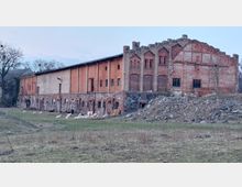 Altes, großes Backsteingebäude mit teils eingestürztem Dach und dekorativen Ziegelmustern, umgeben von einer Wiese, auf der Schafe grasen. Der untere Bereich besteht aus Natursteinmauerwerk, während das Umfeld von Bäumen und unebenem Gelände geprägt ist.
