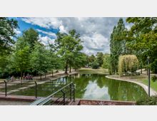 Parkanlage mit einem kleinen, bepflasterten Teich, umgeben von Bäumen und gepflasterten Wegen. Im Vordergrund sind Sitzbänke und ein Geländer sichtbar, während im Hintergrund Spaziergänger entlang der Wege zu sehen sind.