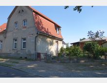 Ein zweigeschossiges Wohnhaus mit hellgrauer Fassade und roten Dachziegeln, teils mit abgebröckeltem Putz, steht an einer gepflasterten Straße. Im Vordergrund befindet sich ein kleiner umzäunter Garten mit grünen Büschen und Bäumen.