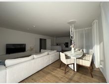 Ein modernes Wohnzimmer mit offener Küche, eingerichtet mit einer hellen Eckcouch, einem Fernseher auf einem niedrigen Schrank und einem runden Esstisch mit vier Stühlen. Der Raum hat einen Boden aus Holzoptik, weiße Wände und Vorhänge, die das Tageslicht weich einlassen.