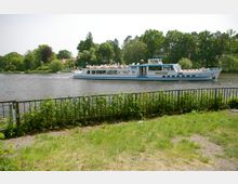 Ein weiß-blaues Ausflugsschiff fährt auf einem Fluss, umgeben von grünen Bäumen und Uferlandschaft. Im Vordergrund ist ein Zaun mit begrüntem Boden zu sehen.