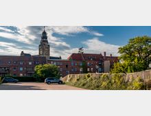 Weitläufiger Parkplatz mit parkenden Autos im Vordergrund, dahinter eine Reihe von roten Backsteinhäusern mit roten Ziegeldächern und Balkonen. Im Hintergrund ragt ein Kirchturm mit Uhr und spitzer Turmspitze in die Höhe, umgeben von Bäumen und einer gepflegten Hecke an der Seite.