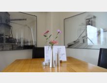 Ein minimalistischer Holztisch in einem modernen Innenraum, dekoriert mit einer schmalen Vase mit Blumen, einer Kerze und einer Getränkekarte. Im Hintergrund hängen zwei großformatige, monochrome Bilder mit architektonischen Motiven an einer neutralen Wand.