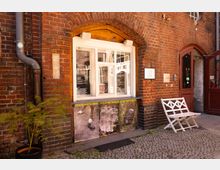 Eingangsnische eines Backsteingebäudes mit großem, weiß gerahmtem Kassenfenster unter einem Rundbogen und einem angrenzenden braunen Türportal. Vor der Fassade stehen ein weißer Klapptisch mit zwei Stühlen auf Kopfsteinpflaster, daneben ein Topf mit kleinem Baum sowie sichtbare Fallrohre und Hinweisschilder an der Wand.