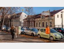 Straßenszene mit parkenden Autos und einer Fußgängerin auf dem Bürgersteig, umgeben von kahlen Bäumen. Im Hintergrund stehen hellfarbene Wohngebäude mit historischen Fassaden, Straßenschilder und ein orangefarbener Mülleimer sind sichtbar.
