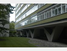 Außenansicht eines modernen Wohngebäudes mit langen Fensterreihen und olivgrüner Fassade. Das Gebäude steht auf markanten V-förmigen Betonstützen, umgeben von einer Rasenfläche mit Bäumen im Hintergrund.