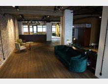 Großzügiger, modern gestalteter Innenraum mit freigelegtem Mauerwerk, Holzboden und Beton-Elementen. Der Raum ist mit einem grünen Sofa, Sesseln, kleinen Tischen und einer Bar im Hintergrund ausgestattet, während helle Fenster und gelbe Vorhänge für Lichtakzente sorgen.