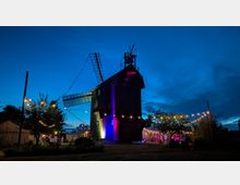 Eine große, hölzerne Windmühle steht in der Abenddämmerung auf einem offenen Gelände, mit langen Flügeln und einem kleinen Aufbau auf dem Dach. Lichterketten und Zelte mit Beleuchtung umgeben die Mühle, Bäume und Nebengebäude rahmen die Szene ein.