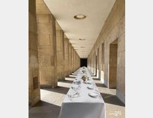 Langer, überdachter Säulengang aus beige-gelbem Stein mit geraden Linien, in dem ein festlich gedeckter Tisch mit weißer Tischdecke, Tellern, Gläsern und Blumendekoration in der Mitte steht. Die Decke ist mit runden Deckenleuchten versehen, und die rechteckigen Wandöffnungen lassen Licht in den Gang.