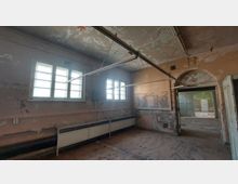 Ein verlassener, stark verfallener Raum mit abblätternder Farbe an den Wänden und der Decke. Zwei große Fenster lassen Licht herein, darunter sind alte Heizkörper zu sehen. Eine offene Tür führt in einen weiteren, ebenfalls heruntergekommenen Raum.