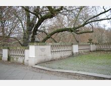 Steinbalustrade entlang eines Gehwegs mit moosbewachsenen, kahlen Bäumen im Hintergrund. Der Boden grenzt teils an Kopfsteinpflaster, teils an gepflastertes Gras; an der Balustrade ist eine Beschriftung angebracht.