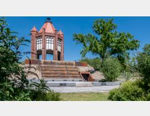 Backsteinpavillon mit spitzem Dach im neugotischen Stil, umgeben von Grünflächen und Bäumen. Im Vordergrund ein gestufter Wasserspielbrunnen mit flachem Steinbecken und kleinen Treppen.