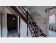 Alt-Text: Treppenhaus in einem alten, verlassenen Gebäude. Die Wände sind abblätternd, die Holztreppe wirkt abgenutzt, und durch ein Fenster im Hintergrund fällt Tageslicht in den Raum.