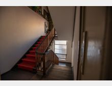 Treppenhaus in einem Wohn- oder Bürogebäude mit rotem Teppichläufer auf den Stufen, Holzgeländern und weißen Wänden. Im Hintergrund befindet sich ein großes Fenster mit Blick auf eine gläserne Außenfront, sowie einige Bilder an der Wand.