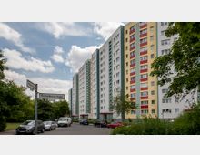 Mehrstöckige Wohnblocks mit bunten Fassaden in Gelb, Weiß und Rot, umgeben von Bäumen und geparkten Autos an einer ruhigen Straße. Straßenschilder mit den Namen „Fichtelbergstraße“ und „Pekrunstraße“ sind im Vordergrund sichtbar.