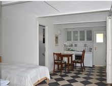 Ein helles Zimmer mit offener Gestaltung, das eine kleine Küchenzeile, einen Essbereich mit Holztisch und zwei Stühlen sowie ein Bett zeigt. Der Boden ist mit schwarz-weiß karierten Fliesen ausgelegt, und im Hintergrund befindet sich eine Tür sowie ein Zugang zu einem kleinen Badezimmer.