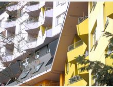 Fassadendetail eines modernen Wohngebäudes mit gelben und weißen Fassadenelementen sowie geometrisch angeordneten Balkonen. Im Vordergrund sind kahle Äste eines Baumes zu sehen, die einen Teil der Fassade verdecken.