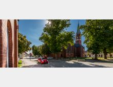 Straßenszene mit einer Backsteinkirche im neugotischen Stil mit Turm und Spitzdach im Hintergrund, umgeben von Bäumen. Im Vordergrund befinden sich ein gepflasterter Gehweg, parkende Autos und ein rotes Fahrzeug am Straßenrand.