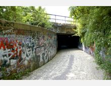 Ein Fußweg führt durch eine Unterführung mit Betonwänden, die vollständig mit Graffiti bedeckt sind, umgeben von dichtem Grün und Kletterpflanzen; oberhalb der Unterführung verläuft eine Brücke mit Geländer.