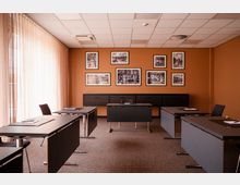 Konferenzraum mit orangefarbenen Wänden, rechteckigen Tischen und schwarzen Stühlen. An der Wand hängen mehrere schwarz-weiße gerahmte Bilder, und der Raum wird von einem großen Fenster mit transparenten Vorhängen beleuchtet.