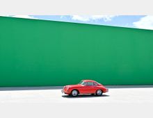 Ein rotes, klassisches Auto steht auf einer hellen Betonfläche vor einer großen, grünen Wand unter einem blauen Himmel mit vereinzelten weißen Wolken.