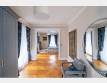 Ein elegant eingerichteter Innenraum mit Holzboden im Fischgrätmuster, einem großen Spiegel an der Wand und langen Vorhängen aus blauem Stoff vor weißen Fenstern. Im Hintergrund ist ein Schlafzimmer mit einem Bett und Wandbildern sichtbar.