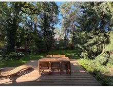 Holzterrasse mit Esstisch, mehreren Stühlen und einer geschwungenen Holzliege, die in einen großen, grünen Garten führt. Der Garten ist von hohen Laub- und Nadelbäumen umgeben, mit Rasenfläche, Sträuchern und einem teilweise sichtbaren Haus im Hintergrund.