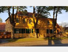 Gelbes zweigeschossiges Wohnhaus mit grün gestrichenen Fensterläden, umgeben von mehreren Bäumen, die Schatten auf die Fassade werfen. Im Vordergrund befindet sich eine gepflasterte Straße, der Hauseingang führt über einen schmalen Weg durch einen kleinen Vorgarten.