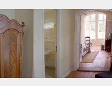 Innenraum eines Wohnbereichs mit Holzboden, einem antiken Holzschrank, einem angrenzenden Badezimmer mit weißen Fliesen und einem kleinen Waschbecken. Im Hintergrund ein Raum mit einem Metallbettgestell, einem Stuhl und großen Fenstern, die viel Tageslicht hereinlassen.