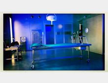 Ein steriler Autopsieraum mit einem beleuchteten Obduktionstisch in der Mitte, einer metallenen Arbeitsstation mit Lampe und Büchern links sowie einer Wand mit Türen und Kühleinheit im Hintergrund. Die Beleuchtung ist in kühlen Blautönen gehalten, und ein medizinisches Personal ist rechts im Bild zu sehen.