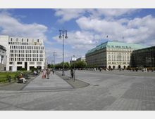Weitläufiger Platz mit gepflastertem Boden in einer städtischen Umgebung. Links ein modernes Gebäude mit schlichten Fensterreihen und einer kleinen Grünanlage, rechts ein historisches Gebäude mit grünem Dach (Hotel Adlon) und ein zeitgenössisches verglastes Bauwerk; Menschen spazieren und sitzen auf Bänken unter einem blauen Himmel mit Wolken.